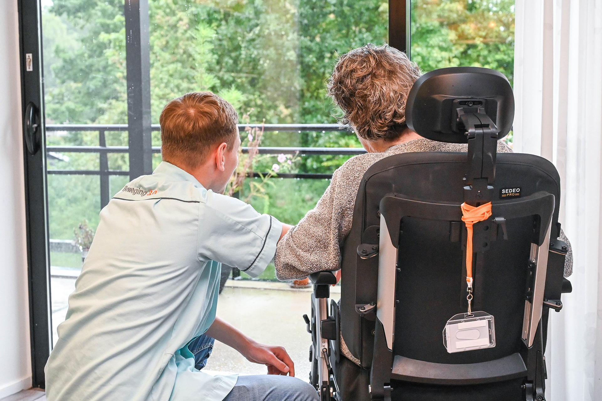 Een Less2Care verzorger knielt naast een oudere volwassene in een rolstoel. Beiden kijken uit een groot raam naar het groen. De scène straalt steun en gezelschap uit in een rustige omgeving binnenshuis.