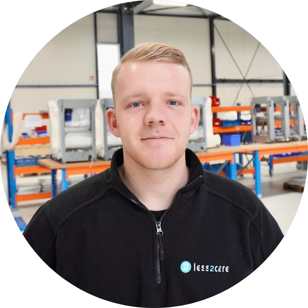 Een jonge man met kort blond haar staat binnen, gekleed in een zwart jasje met het less2care-logo. Achter hem staan werkbanken, rekken en industriële apparatuur in een helder verlichte werkplaats of fabrieksomgeving.